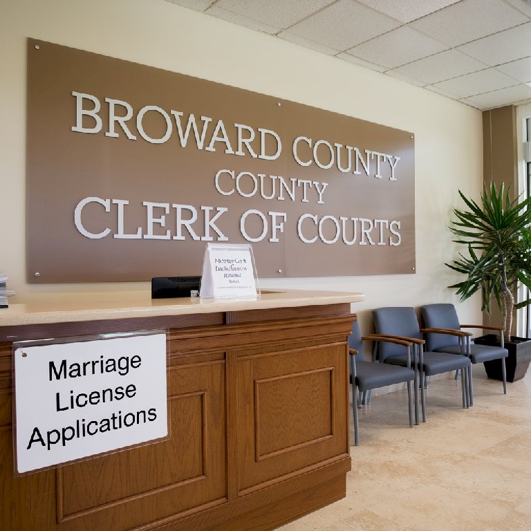 A photo of a Broward County Marriage License office. There is a wooden counter with a sign that says "Marriage License Applications". On the wall behind the counter, there's a large beige sign with white text that says "Broward County Clerk of Courts". There are a few chairs for waiting and a potted plant beside the wall. The floor is made of beige tiles. The lighting is soft.