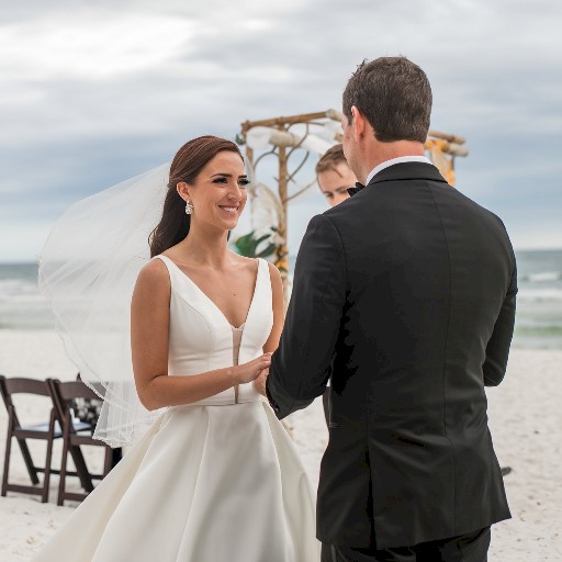 A photo of a wedding ceremony taking place in Orange County, Florida. The bride is wearing a white dress and a veil, while the groom is wearing a black tuxedo. They are standing on a sandy beach. The background contains a few chairs and a decorative arch. The sky is overcast.